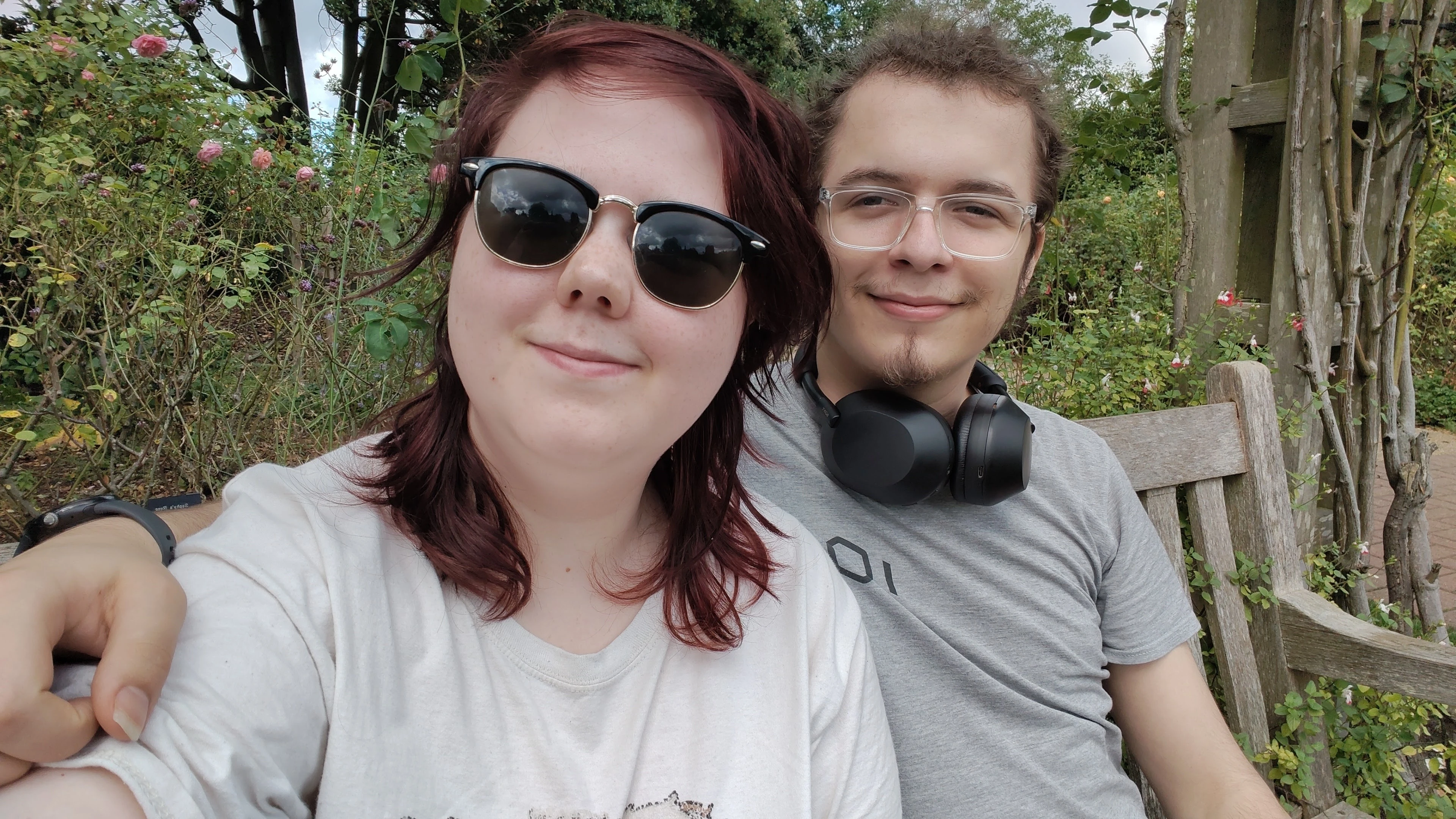 Close up photo of two smiling individuals, the one on the left has red hair and is wearing sunglasses, the one on the right has their arm around the other and has clear glasses with their hair tied back